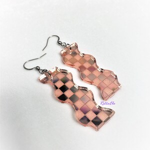 May include: A pair of rose gold-coloured, wavy-shaped earrings with a checkerboard pattern. The earrings have a metallic sheen and are suspended from dark grey ear wires. The design incorporates black and pink squares.