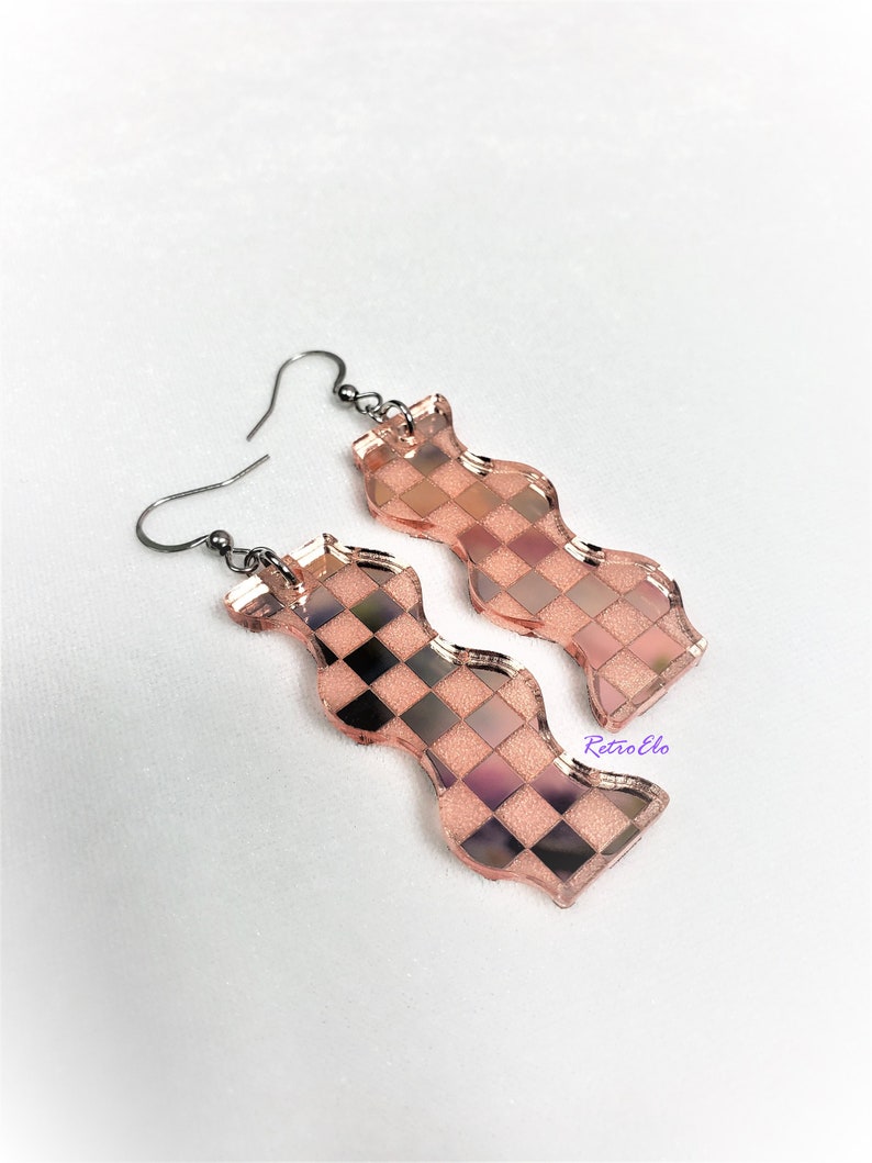 May include: A pair of rose gold-coloured, wavy-shaped earrings with a checkerboard pattern. The earrings have a metallic sheen and are suspended from dark grey ear wires. The design incorporates black and pink squares.