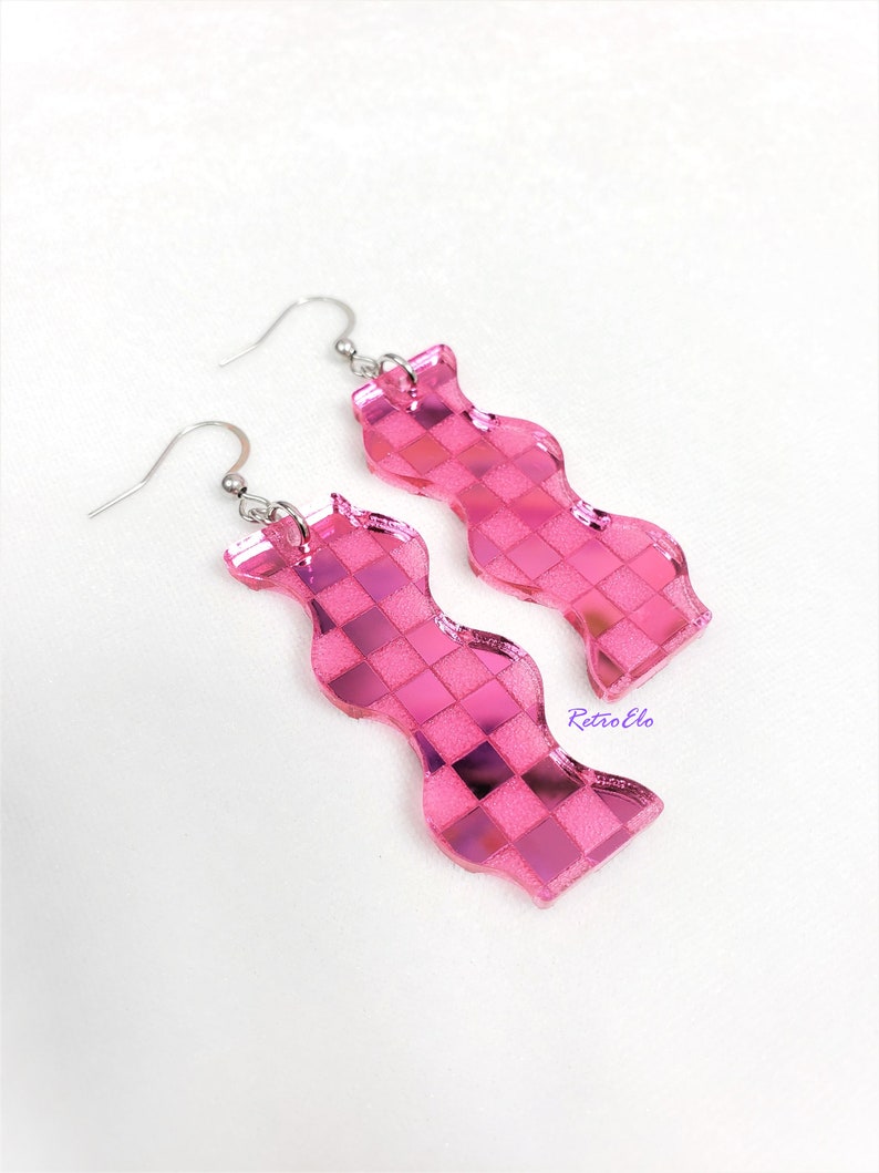 May include: A pair of pink, wavy earrings with a checkered design. The earrings have a shiny, reflective surface and are attached to silver-coloured ear wires. The word "RetroElo" is printed on the right earring.