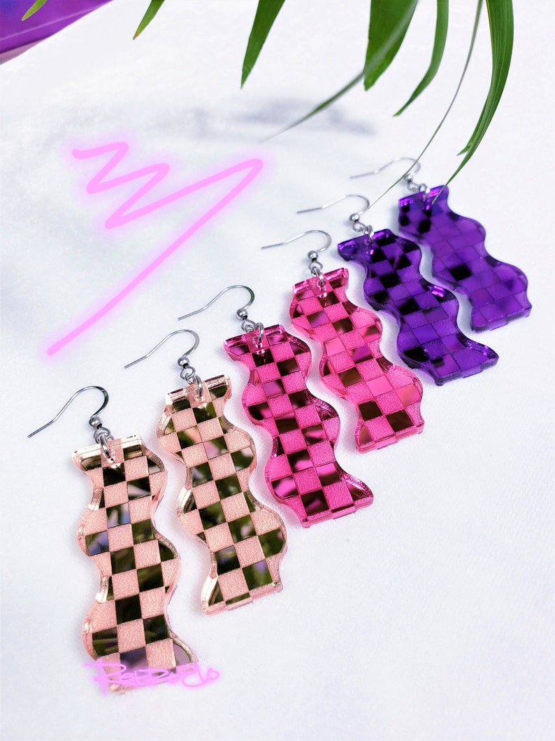 May include: A selection of colourful, wavy-shaped earrings in shades of pink, purple, and peach. Each earring has a checkered pattern and a silver hook. The earrings are displayed on a white surface with a neon pink squiggle.