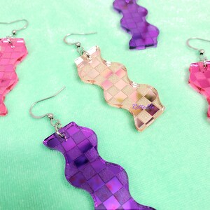 May include: Wavy earrings in pink, peach, and purple, featuring a checkered pattern. The earrings have silver-coloured hooks. The earrings are displayed on a light green surface. The design is geometric.