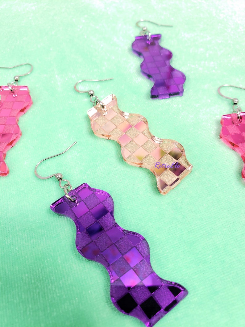 May include: Wavy earrings in pink, peach, and purple, featuring a checkered pattern. The earrings have silver-coloured hooks. The earrings are displayed on a light green surface. The design is geometric.