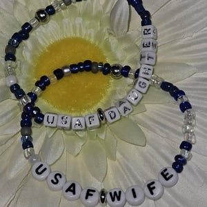 May include: Two beaded bracelets with white letter beads spelling out "USAF WIFE" and "USAF DAUGHTER". The bracelets are blue and silver.