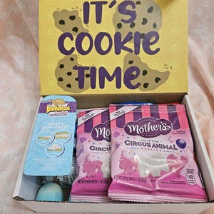 May include: A white box with a yellow lid that says "It's Cookie Time" with a chocolate chip cookie design. Inside the box are two pink bags of Mother's Circus Animal Cookies, each weighing 1 ounce, and a blue package of Dunkaroos.