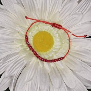 May include: A red beaded bracelet with an adjustable sliding knot closure. The bracelet is displayed on a white flower with a yellow center. The beads are small and faceted, reflecting light.