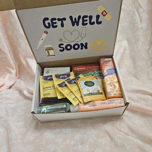 May include: A white cardboard box with the words "Get Well Soon" on the lid. The box is filled with various items, including tea bags, hand sanitizer, and a lemon ginger drink mix.