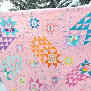 May include: A pink quilt with a repeating pattern of stars, triangles, and squares. The quilt features various colours, including teal, orange, purple, and yellow. Some squares have images of a girl, a rainbow, and a camper.