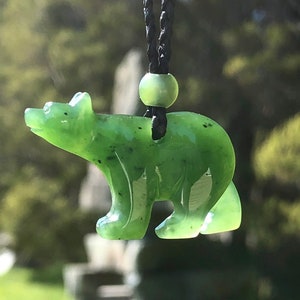 May include: A green jade bear pendant carved in a standing pose, suspended from a black cord. The bear has a smooth, polished surface with dark flecks. A green bead accents the cord above the bear.