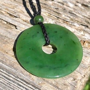 May include: A green jade pendant in the shape of a donut, hanging from a black cord necklace. The pendant has a smooth, polished surface with dark green speckles.