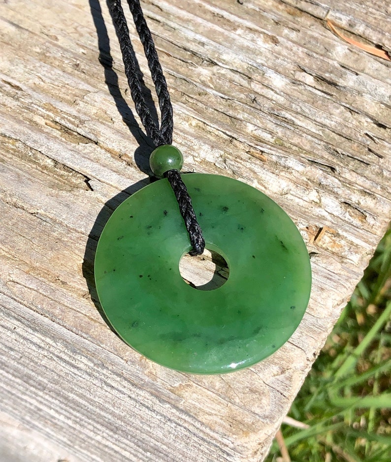 May include: A green jade pendant in the shape of a donut, hanging from a black cord necklace. The pendant has a smooth, polished surface and a hole in the center.