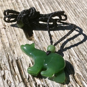 May include: A green jade bear pendant on a black cord. The bear is carved in a standing pose, with visible legs and a rounded body. The cord has a small jade bead and is knotted at the top. The pendant is set against a weathered wooden surface.