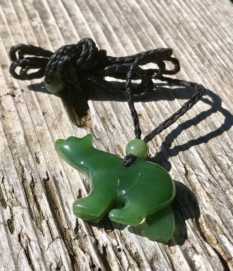 May include: A green jade bear pendant on a black cord. The bear is carved in a standing pose, with visible legs and a rounded body. The cord has a small jade bead and is knotted at the top. The pendant is set against a weathered wooden surface.