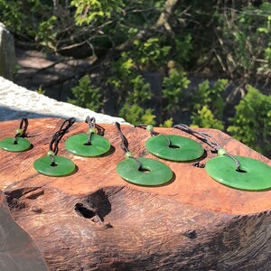 May include: A set of six green jade pendants in various sizes, each with a hole in the center and strung on a black cord. The pendants are arranged on a wooden surface.
