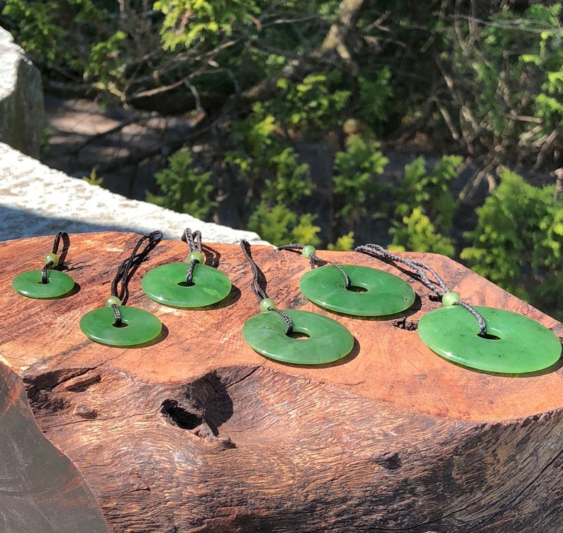 May include: A set of six green jade pendants in various sizes, each with a hole in the center and strung on a black cord. The pendants are arranged on a wooden surface.