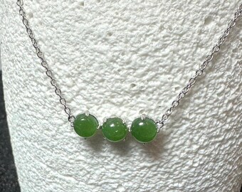 May include: A delicate necklace featuring three round, vibrant green jade beads. The beads are strung on a fine silver chain. The necklace is displayed against a textured white background, highlighting the jewelry's simple elegance.