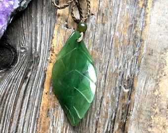May include: A green jade leaf pendant with carved details hangs from a brown cord necklace. The leaf-shaped pendant is a vibrant green color and has a polished finish. The necklace is displayed on a rustic wooden surface.