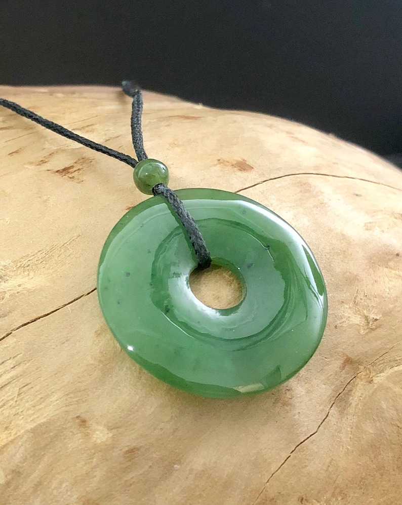 May include: A green jade pendant in the shape of a ring, hanging from a black cord. The pendant is smooth and polished, with a hole in the center.