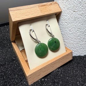 May include: A pair of green jade earrings with silver hooks, presented in an open wooden box. The earrings are circular and polished, with a dark green hue. The box is made of light-coloured wood and the earrings are displayed against a cream background.
