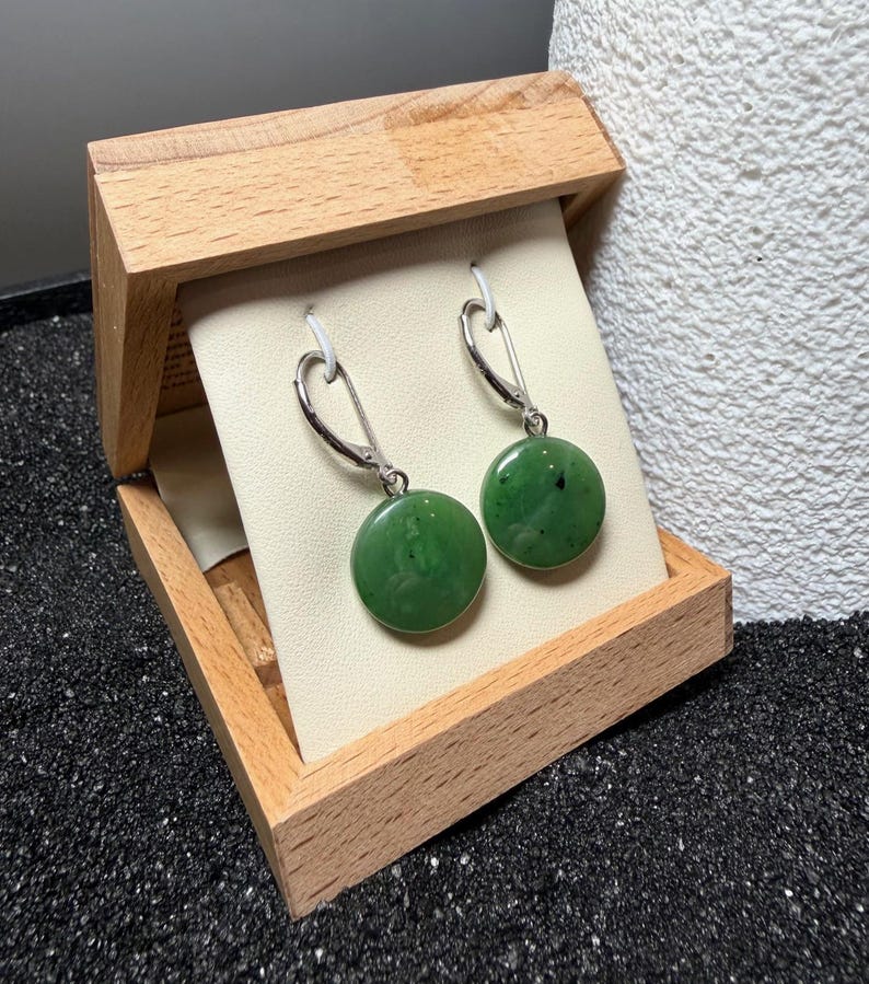 May include: A pair of green jade earrings with silver hooks, presented in an open wooden box. The earrings are circular and polished, with a dark green hue. The box is made of light-coloured wood and the earrings are displayed against a cream background.