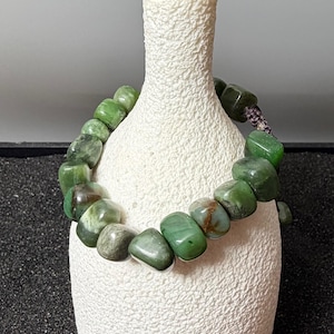 May include: A green stone bracelet featuring rounded, irregular beads. The beads are various shades of green with some brown inclusions. The bracelet is strung on a dark cord and is displayed on a white textured surface.