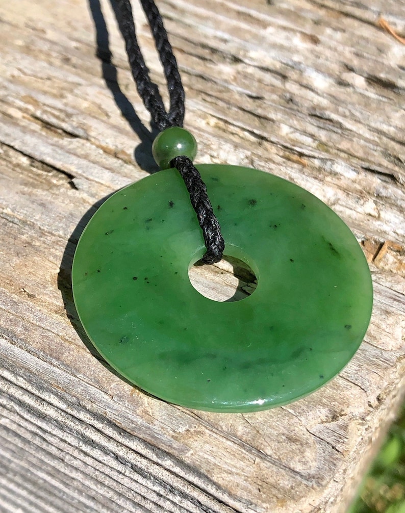 May include: A green jade pendant in the shape of a donut, hanging from a black cord necklace. The pendant has a smooth, polished surface with dark green speckles.