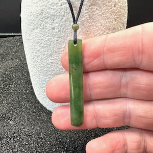 May include: A green jade pendant necklace with a rectangular bar shape. The pendant hangs from a black cord. The jade has a polished finish and natural variations. The necklace is held against a neutral background.