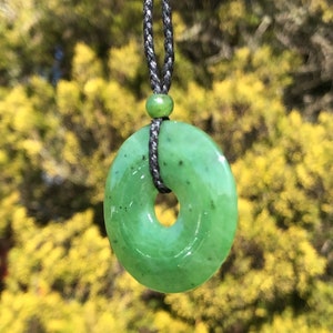 May include: A green jade pendant in the shape of a ring, hanging from a black cord necklace. The pendant has a smooth, polished surface and a subtle swirl pattern.