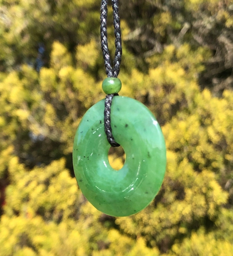 May include: A green jade pendant in the shape of a ring, hanging from a black cord necklace. The pendant has a smooth, polished surface and a subtle swirl pattern.