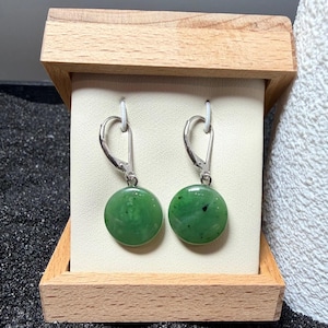 May include: A pair of round, green jade earrings with silver lever-back fastenings. The earrings are presented in a small, light-coloured wooden box with a cream backing.