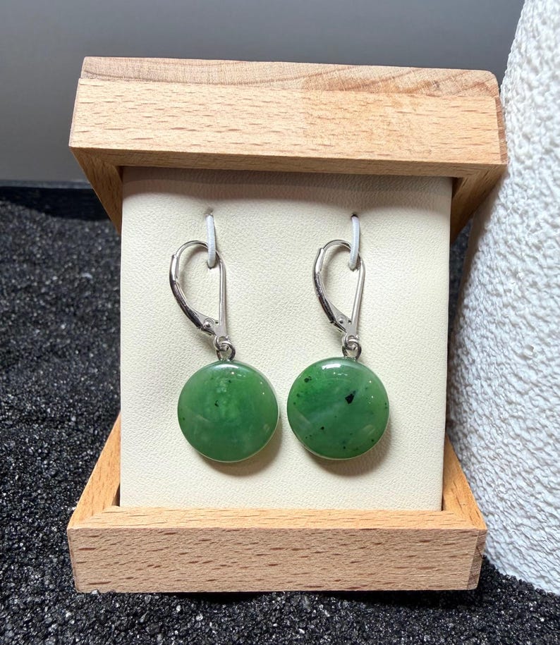 May include: A pair of round, green jade earrings with silver lever-back fastenings. The earrings are presented in a small, light-coloured wooden box with a cream backing.