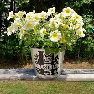 May include: A silver metal planter with a black vinyl decal featuring the letter "H" and the name "HUSKIN" surrounded by floral designs. The planter is filled with white petunias.