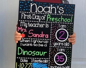 May include: A black chalkboard sign with white, green, pink, and blue text. The sign says "Noah's First Day of Preschool," and includes information about the child's age, teacher, and favorite color. The sign also states the child is 35 inches tall.