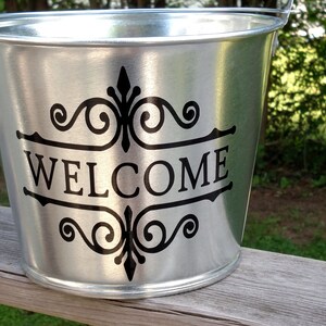 May include: A silver metal bucket with a black vinyl decal that says "WELCOME" with decorative scrollwork.
