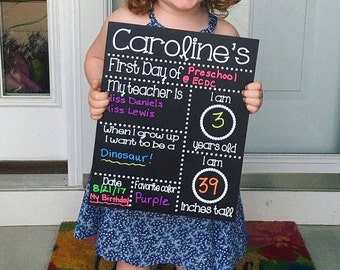 Puede incluir: Un letrero negro de pizarra con texto blanco que anuncia el primer día de preescolar de un niño. El letrero incluye el nombre del niño, su edad, su altura, su color favorito y su cumpleaños. El texto dice: "Caroline's First Day of Preschool @ ECDC. My teacher is Miss Daniels Miss Lewis. I am 3 years old. I am 39 inches tall. Date 8/21/17. Favorite color: Purple. My Birthday: "