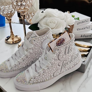 Puede incluir: Zapatillas blancas de caña alta con adornos de perlas y pedrería. Las zapatillas están personalizadas con el texto "Mrs. Jackson".