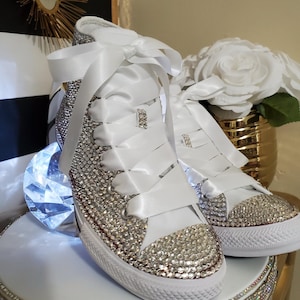 Puede incluir: Zapatillas blancas de caña alta cubiertas de brillantes brillantes con cordones de cinta de satén blanco atados en un lazo.