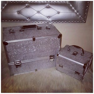 May include: Two silver makeup cases, one large and one small, are covered in sparkling rhinestones. The larger case has a black handle and two latches, while the smaller case has a silver handle and one latch. The cases sit on a fluffy white surface.