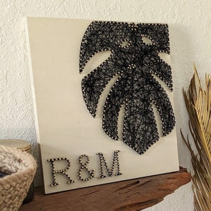 May include: A white wooden sign with a black string art design of a monstera leaf. The initials "R&M" are spelled out below the leaf in gold.