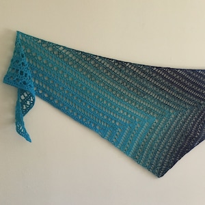 May include: A crocheted shawl with a gradient of blue and teal colors. The shawl is made with a lacy stitch pattern and has a fringed edge.