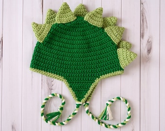 Puede incluir: Sombrero de dinosaurio de crochet verde con un largo cordón trenzado verde y blanco.
