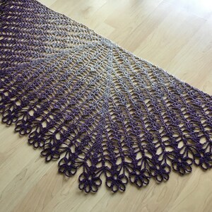 May include: A handmade, open-weave shawl in a gradient of purple to gray. The shawl features a scalloped edge with a floral design. The intricate crochet work creates a delicate and airy appearance. The shawl is displayed on a wooden surface.