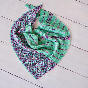 Puede incluir: Un chal de ganchillo verde y morado con un patrón geométrico. El chal está doblado y drapeado sobre una superficie de madera blanca.