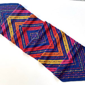 Op de afbeelding: Een lange, gehaakte tafelloper met een geometrisch patroon in tinten blauw, oranje, geel en roze. De loper is gemaakt met een textuur-steek en heeft franjes aan de uiteinden.