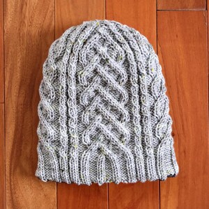 May include: A gray knit beanie with a cable knit design. The hat has a ribbed brim and a textured pattern throughout the crown. The yarn has flecks of yellow and green. The hat is displayed on a wooden surface.