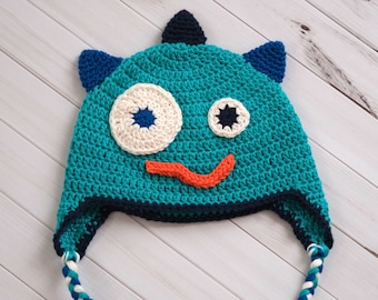 Könnte beinhalten: Eine türkisfarbene Häkelmütze mit einem Monstergesicht-Design. Die Mütze hat ein großes weißes und cremefarbenes Auge, ein kleineres Auge mit einem marineblauen Stern und einen orangefarbenen Mund. Die Mütze hat Ohrenklappen und geflochtene Bänder.
