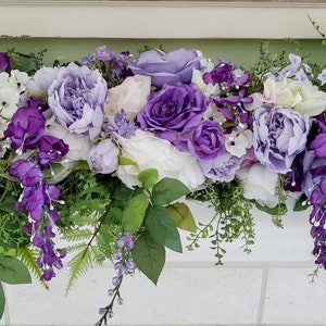 May include: A decorative floral display with a variety of purple, lilac, and white blooms, such as hydrangeas, roses, and wisteria. The arrangement is complemented by green foliage. The flowers are arranged on a white surface.