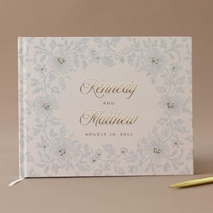 May include: A wedding guest book with a floral design in light blue and white. The names "Kennedy and Matthew" are in gold script, with the date "August 30, 2025" below. A gold pen rests on the side.