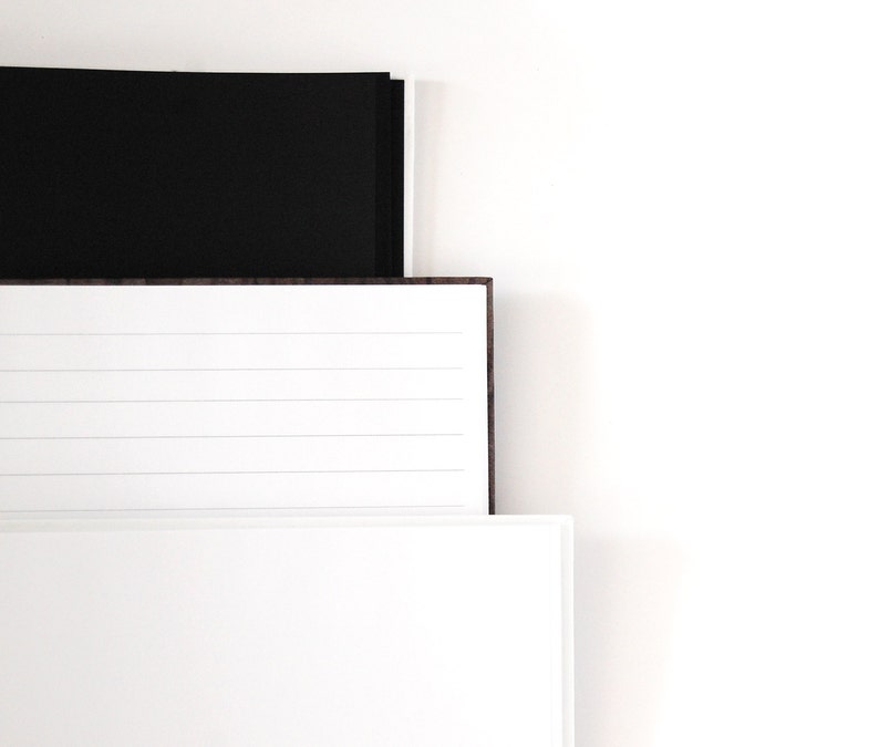 May include: A stack of three notebooks with a minimalist design. The top notebook is black, the middle one has lined pages, and the bottom one is white. The notebooks are arranged against a white background.