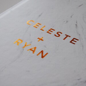 May include: A white marble-patterned book cover with the names "CELESTE + RYAN" in gold foil lettering. The cover has a glossy finish and a subtle, elegant design, suitable for a wedding or special occasion.