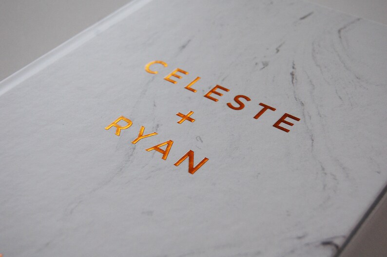 May include: A white marble-patterned book cover with the names "CELESTE + RYAN" in gold foil lettering. The cover has a glossy finish and a subtle, elegant design, suitable for a wedding or special occasion.
